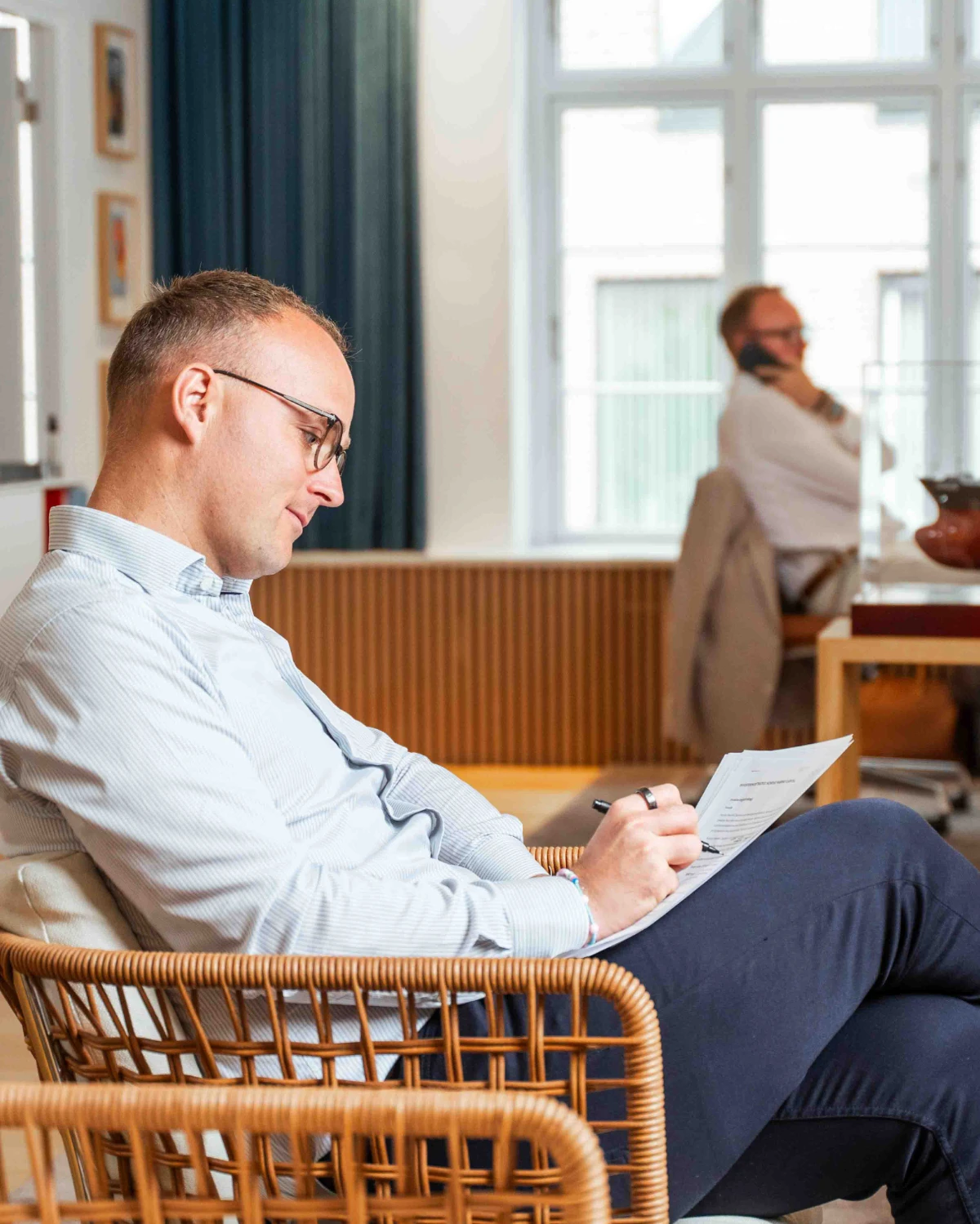 Christian som sidder i en stol og skriver på et dokument, mens Rasmus sidder i baggrunden og snakker i telefon